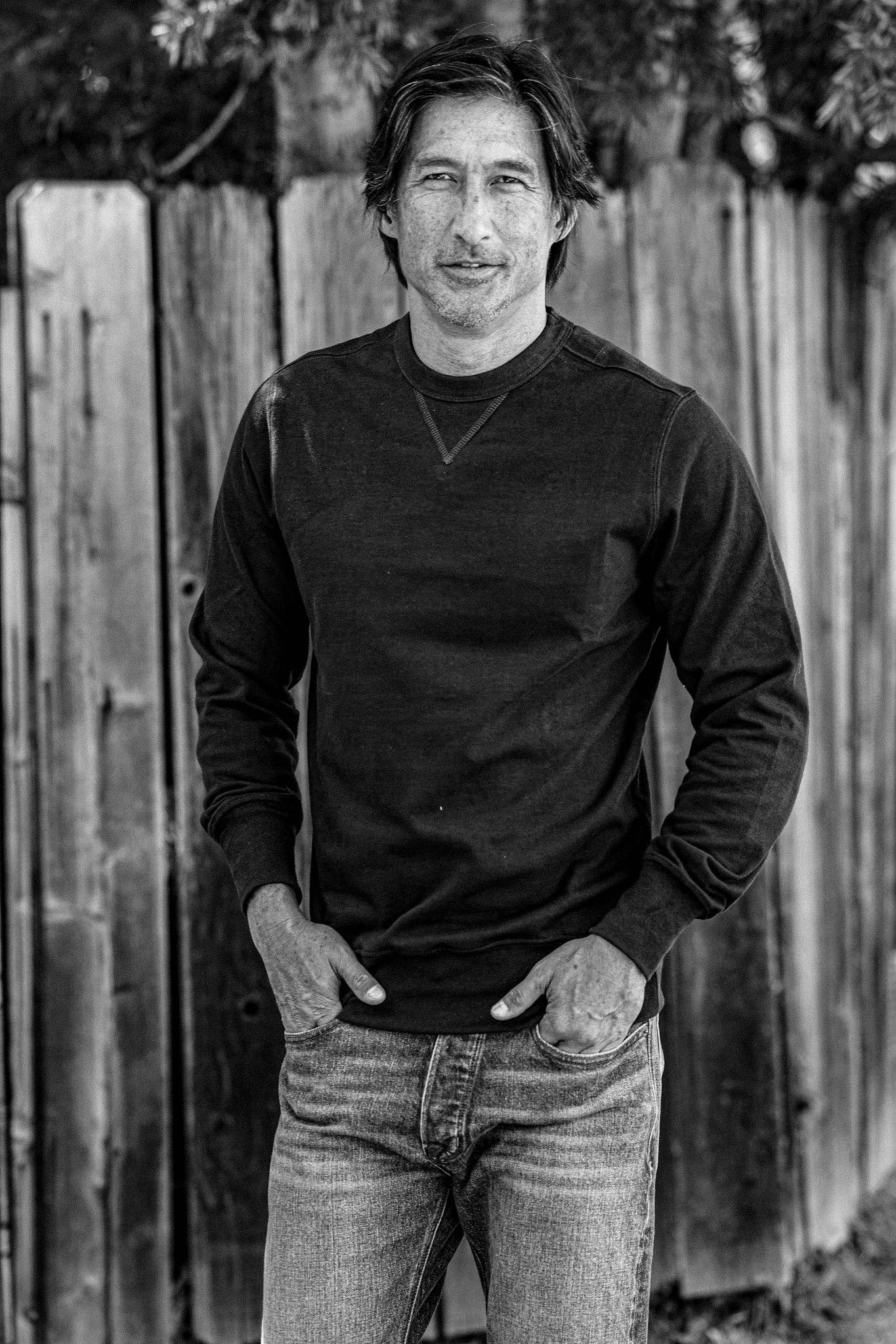 Person wearing a black long-sleeve shirt and jeans standing against a wooden fence.