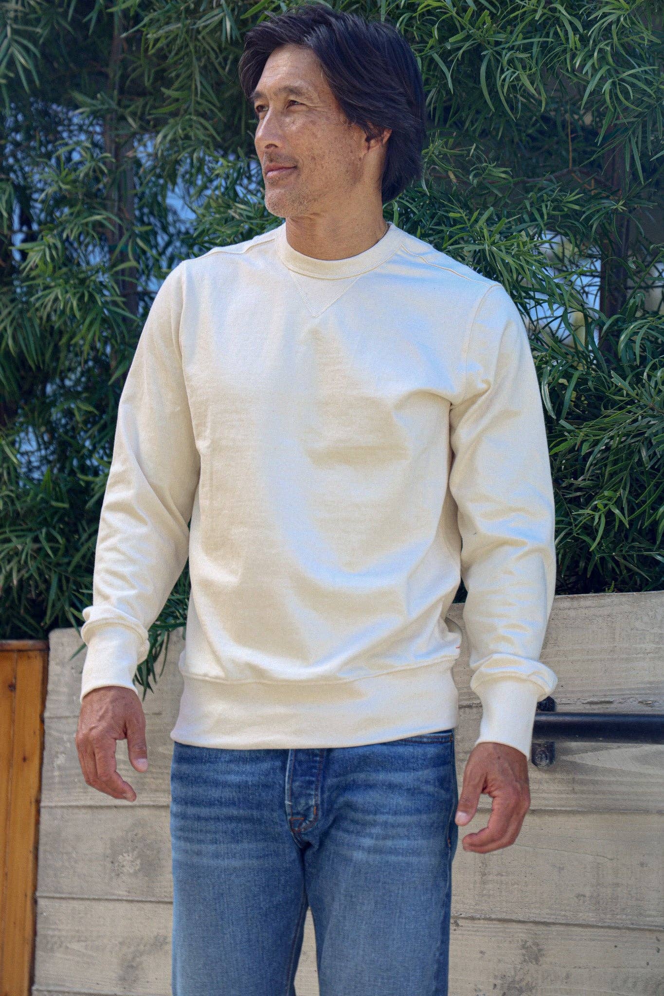 Man wearing a cream sweatshirt and blue jeans standing outdoors with greenery in the background.