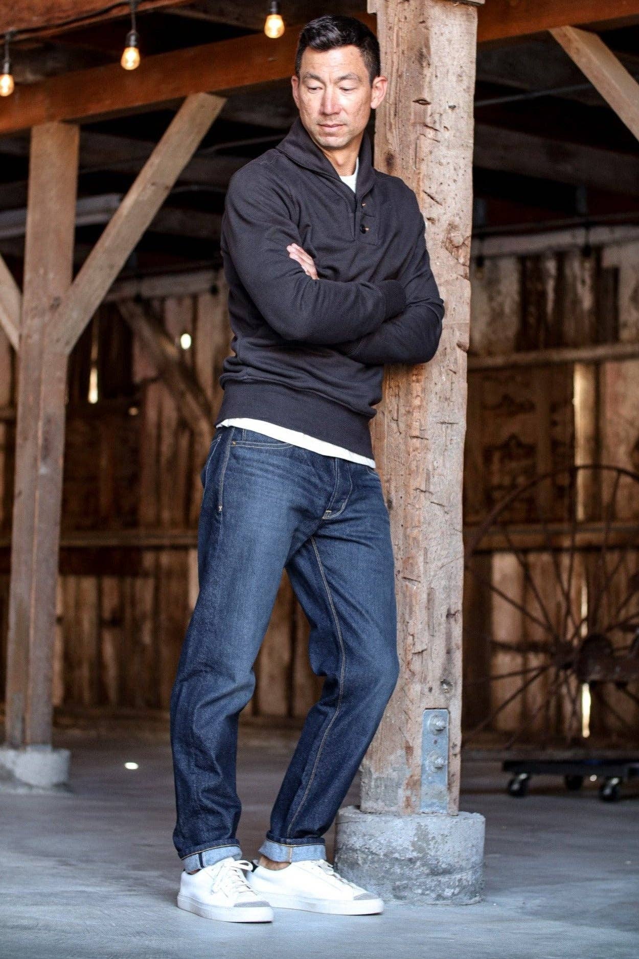 Man wearing a dark top and jeans standing against a wooden post in a rustic setting