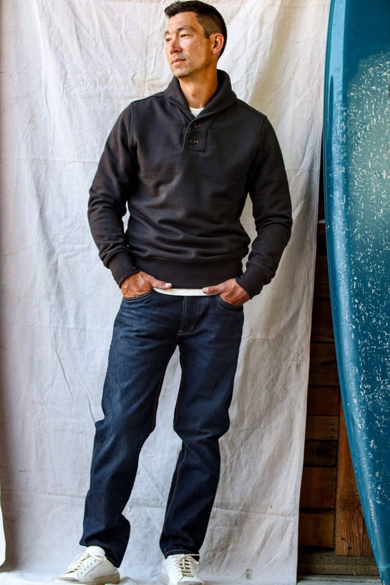 Man wearing a dark gray pullover and blue jeans standing next to a blue surfboard.
