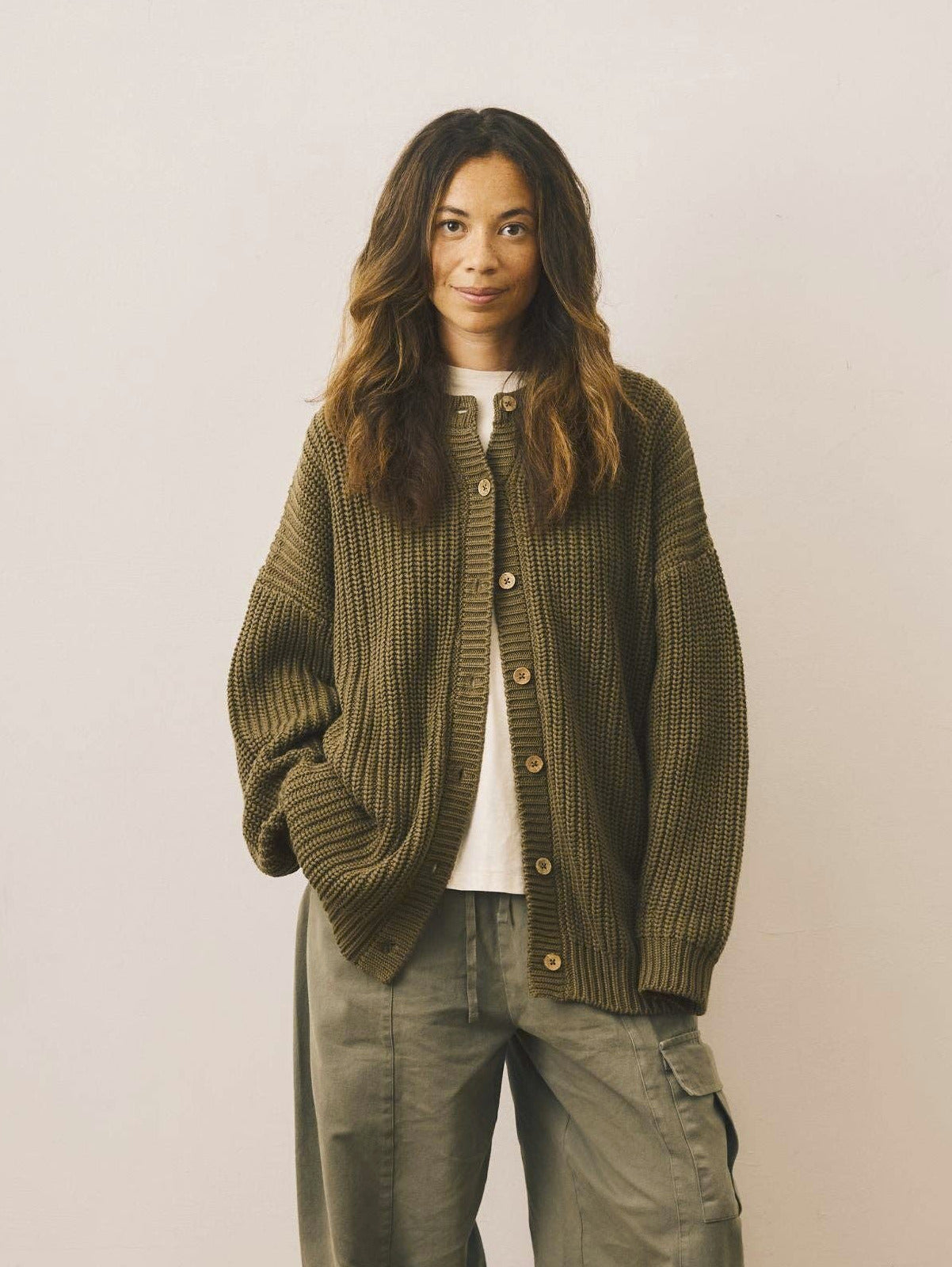 Person wearing a green cardigan and green pants standing on a wooden floor with a white wall background