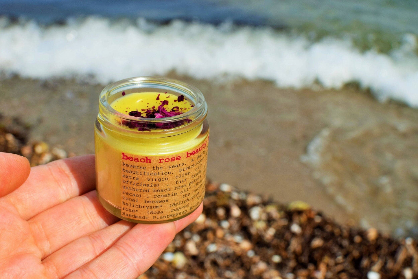 Hand holding a jar of 'beach rose body butter' with ocean waves in the background