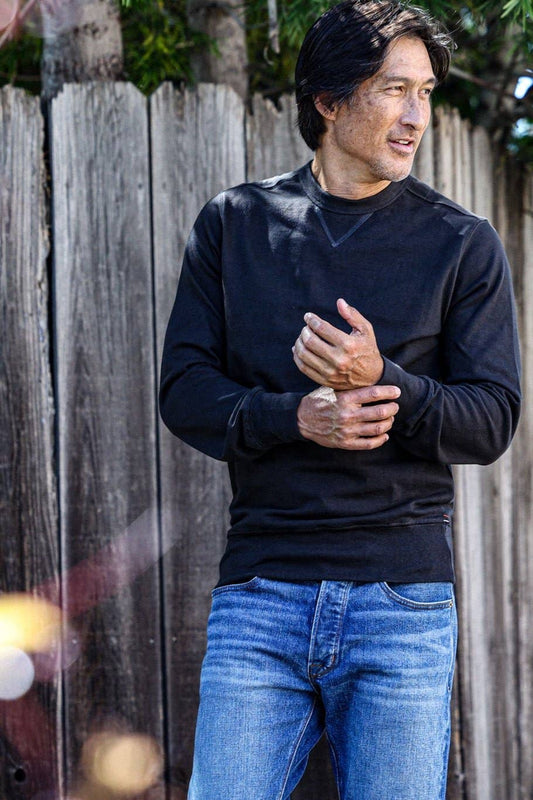 Man wearing a black sweater and blue jeans standing in front of a wooden fence.